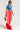 A fashionable person stands confidently wearing a blue "London" crop top, red Modern Jam Track Pant with Tulle featuring white stripes and a high-rise design, and red fluffy shoes. They hold a small leopard print handbag in their right hand. The background is plain white.