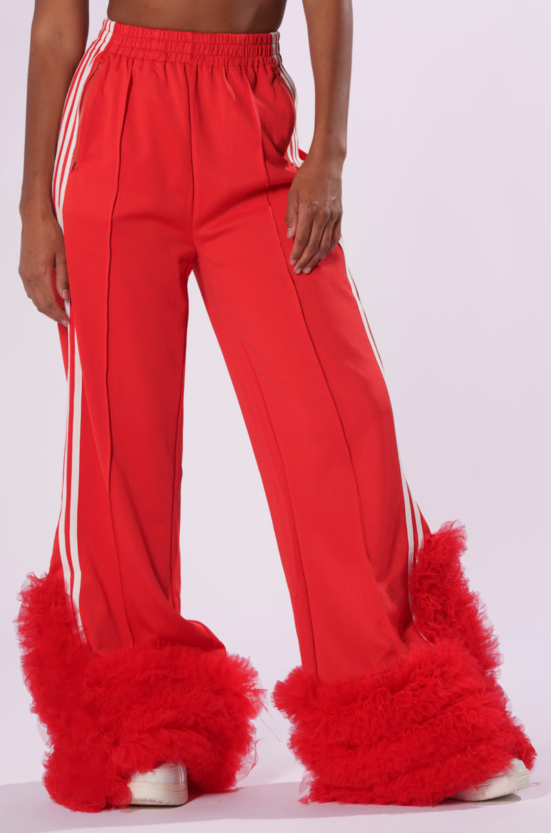 A person is wearing the MODERN JAM TRACK PANT WITH TULLE in a vibrant red color, featuring white side stripes. These high-rise track pants have wide legs embellished with fluffy tulle ruffles at the bottom. The outfit is completed with white shoes, set against a plain, light-colored background.