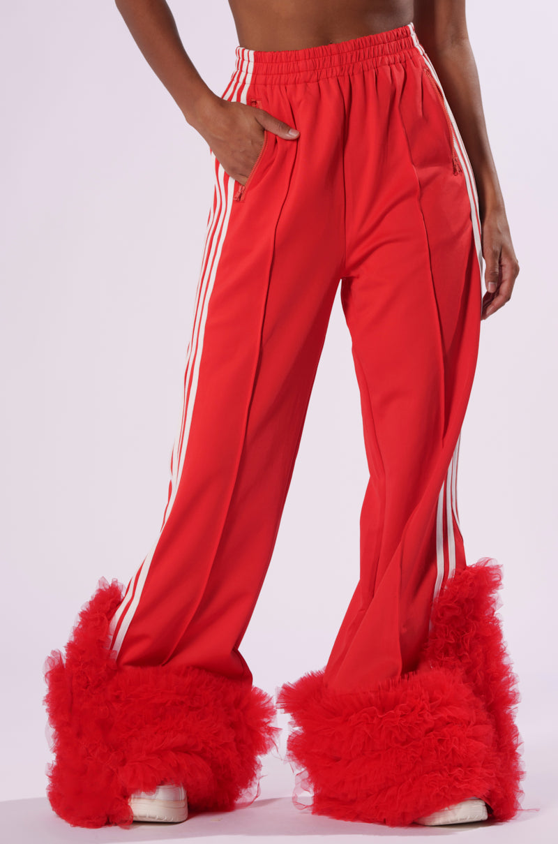 A person is wearing the MODERN JAM TRACK PANT WITH TULLE in vibrant red, adorned with white stripes along the sides. The high-rise track pants are styled with voluminous red tulle ruffles enveloping the lower legs, partially hiding a pair of white athletic shoes. The backdrop is a plain white setting.