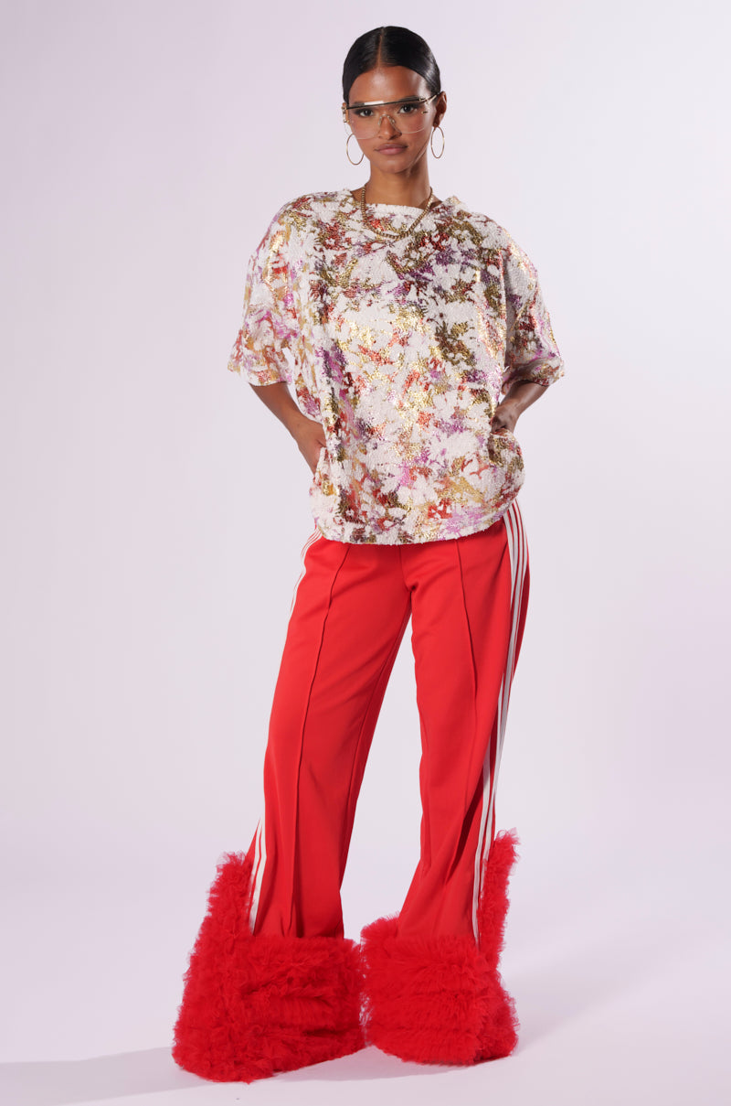 A fashionable woman stands against a plain background wearing clear glasses, a floral-patterned shirt, bright red Modern Jam Track Pant with Tulle featuring white stripes, and matching red fluffy fur-covered boots. She has her hands on her hips, exuding confidence.
