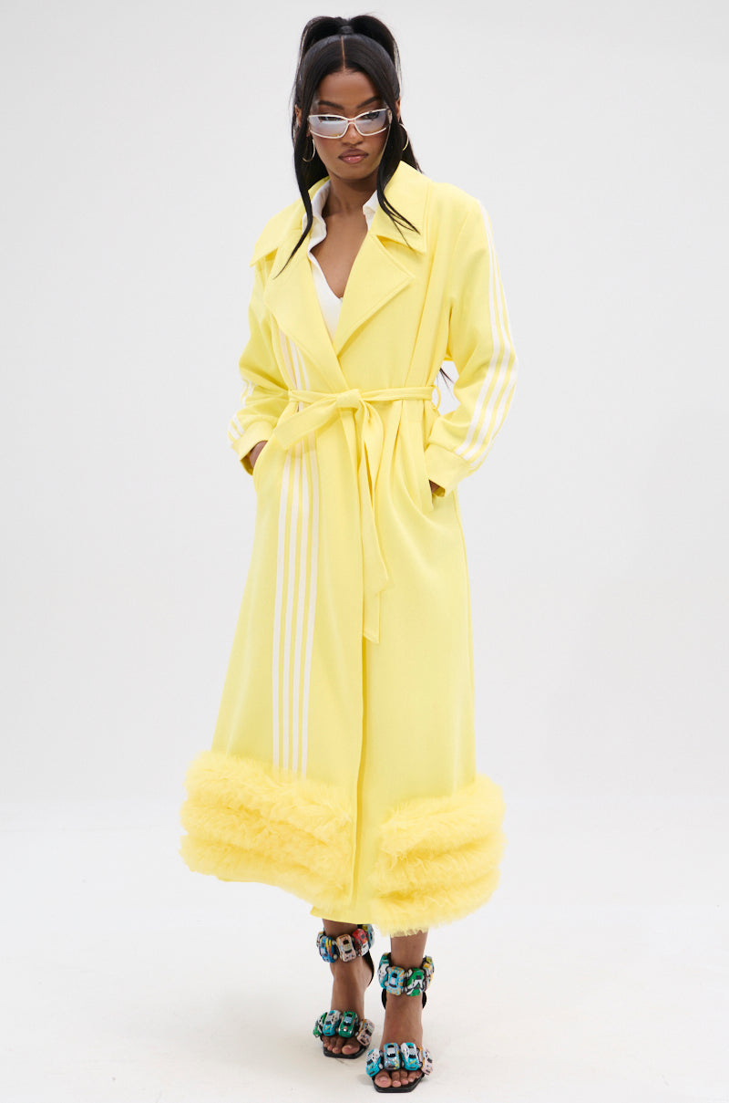 The person stands confidently in the MIMOSA TULLE TRIM TRENCH, a lemon-yellow coat with white stripes and playful tulle trim. An adjustable waist belt enhances their silhouette. Dark-haired and wearing sunglasses, they pair it with colorful block-heeled sandals against a plain white backdrop.