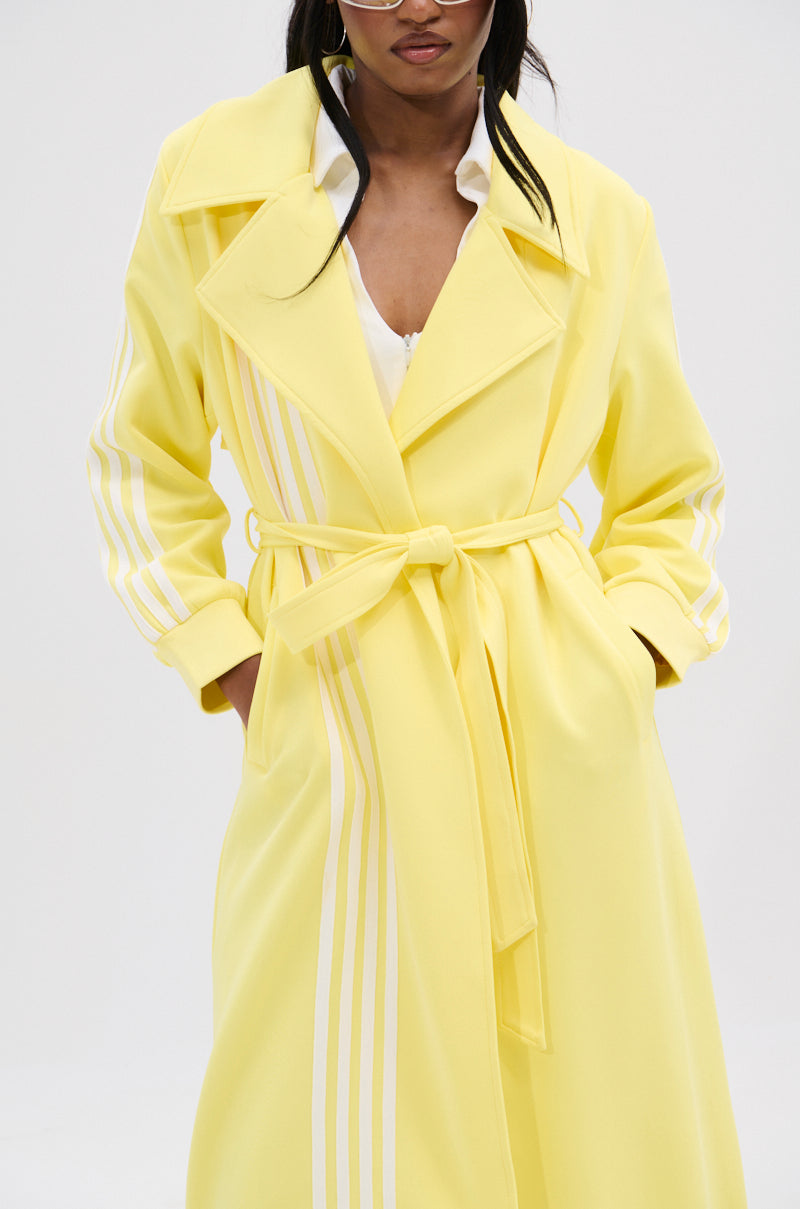 The MIMOSA TULLE TRIM TRENCH, a chic lemon-yellow coat with white sleeve stripes and an adjustable waist belt, is paired with a white top. Complete the sleek style with sunglasses for the perfect look.