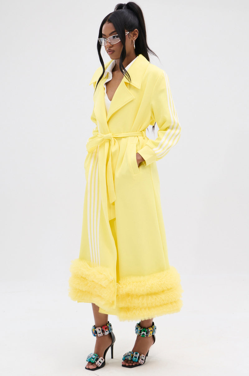 The person is donned in the MIMOSA TULLE TRIM TRENCH, a lemon-yellow coat featuring white sleeve stripes, a fluffy hem trim, and an adjustable waist belt. They're pairing it with vibrant embellished high-heeled sandals and clear futuristic glasses.