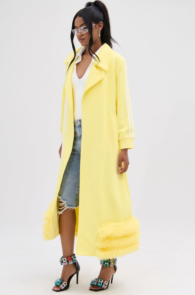 A woman stands against a plain background, wearing glasses and her hair tied back. She poses in the MIMOSA TULLE TRIM TRENCH—a lemon-yellow coat featuring a fuzzy trim at the bottom—paired with distressed denim shorts, a white top underneath, and colorful high-heeled sandals.