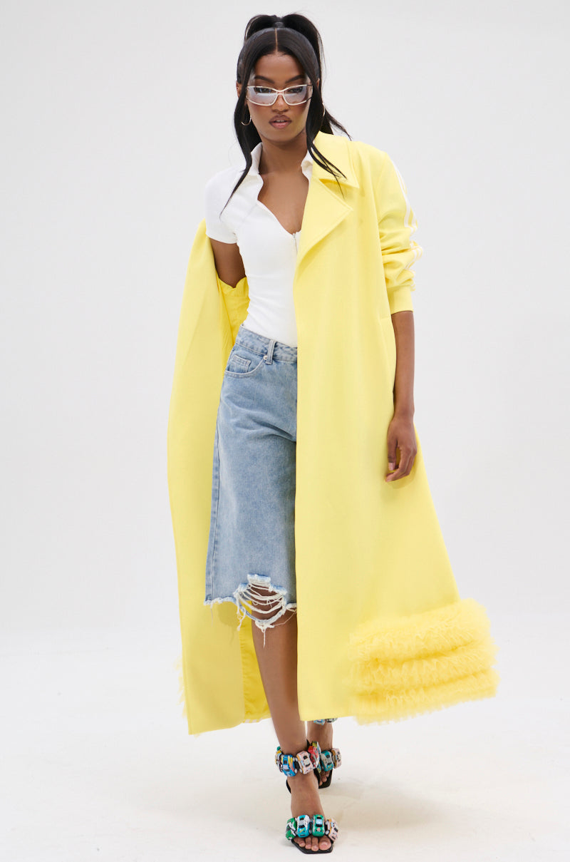A person wears the Mimosa Tulle Trim Trench in lemon-yellow, featuring textured hem details, over a white top and distressed denim shorts. They accessorize with stylish glasses and colorful high-heeled sandals against a plain white backdrop.