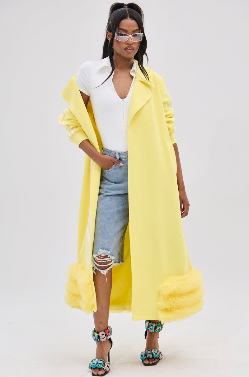 A fashionable individual wears a MIMOSA TULLE TRIM TRENCH in lemon-yellow, complete with an adjustable waist belt and ruffled hem. They pair it with a white top with a unique collar, ripped jeans, colorful high heels, stylish glasses, and let their long dark hair flow against a plain background.