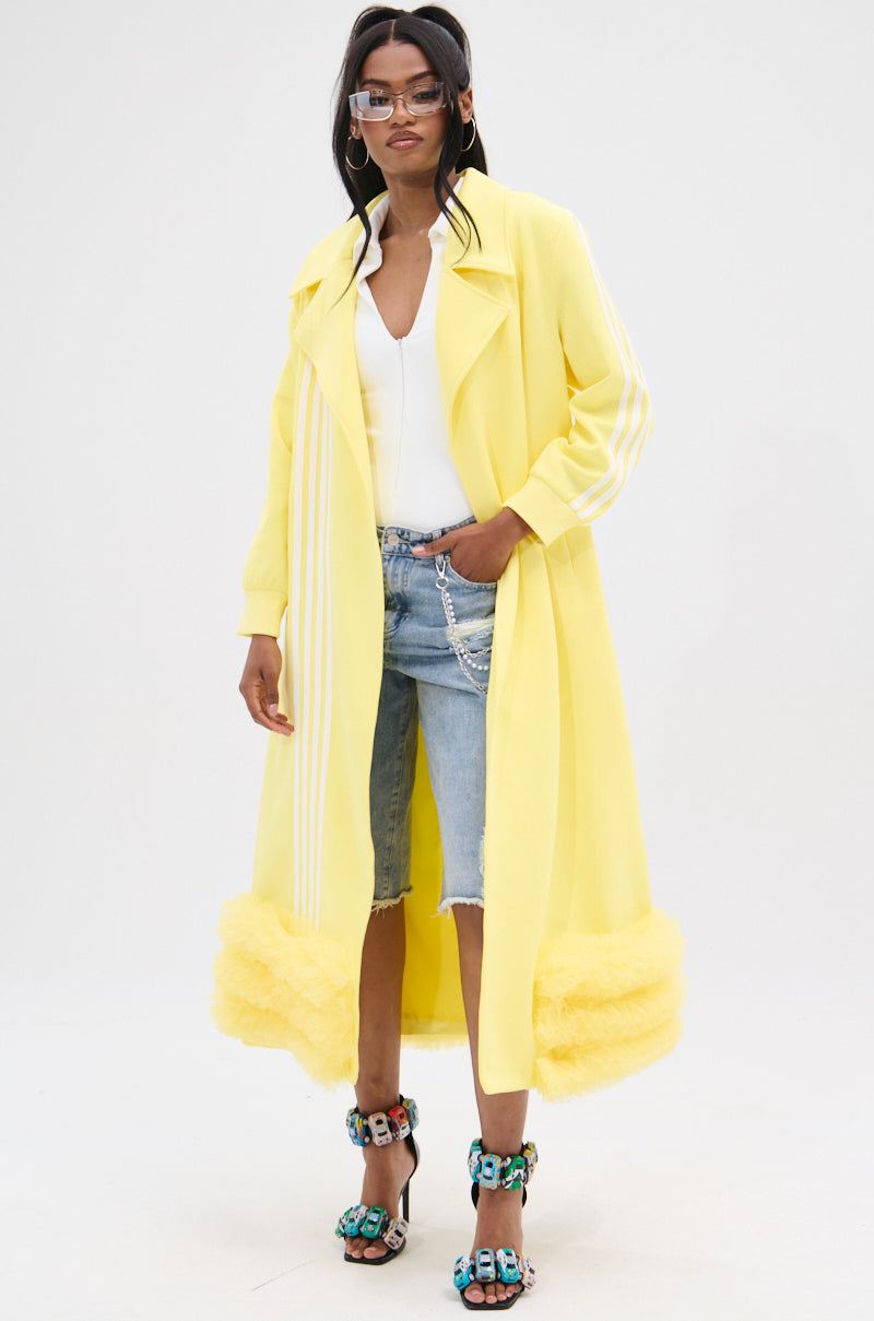 Wearing the MIMOSA TULLE TRIM TRENCH in lemon-yellow with white stripes, a white top, denim shorts, and colorful embellished heels, a person with long dark hair and sunglasses stands confidently against a plain background.