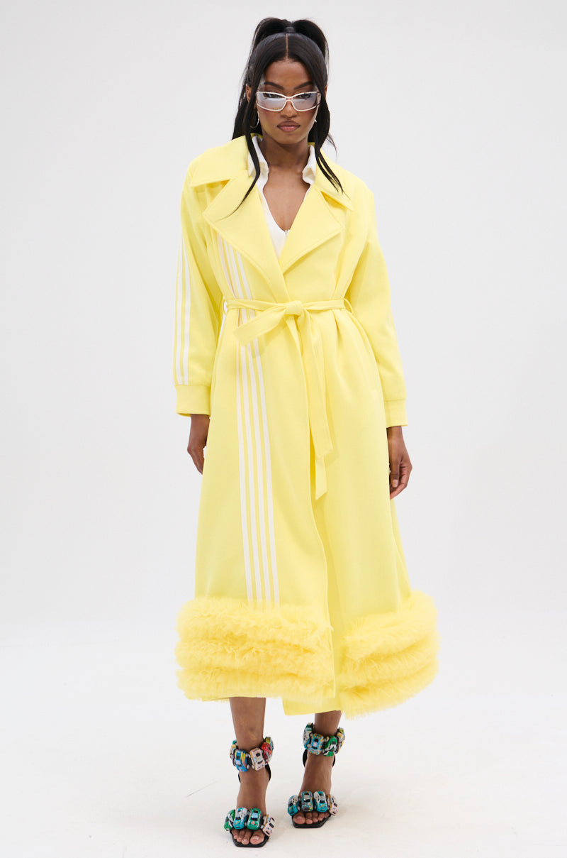 A woman confidently wears the MIMOSA TULLE TRIM TRENCH in lemon-yellow with white stripes. Her clear glasses complement her vibrant high-heeled sandals, and the adjustable waist belt cinches her outfit perfectly against a plain white backdrop.