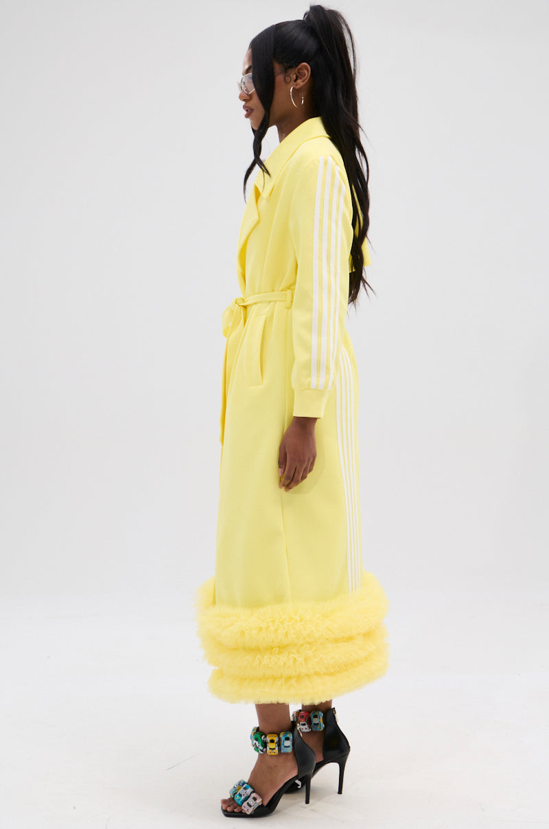 A person with long hair models the MIMOSA TULLE TRIM TRENCH in lemon-yellow, showcasing its adjustable waist belt and tulle trim. They pair it with colorful high heels while standing sideways against a plain background.