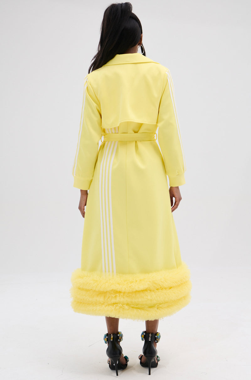 The individual stands turned away, donning the MIMOSA TULLE TRIM TRENCH—a vibrant lemon-yellow coat with white sleeve and back stripes, fluffy yellow hem trim, and an adjustable waist belt. They pair it with black high-heeled shoes featuring decorative embellishments.