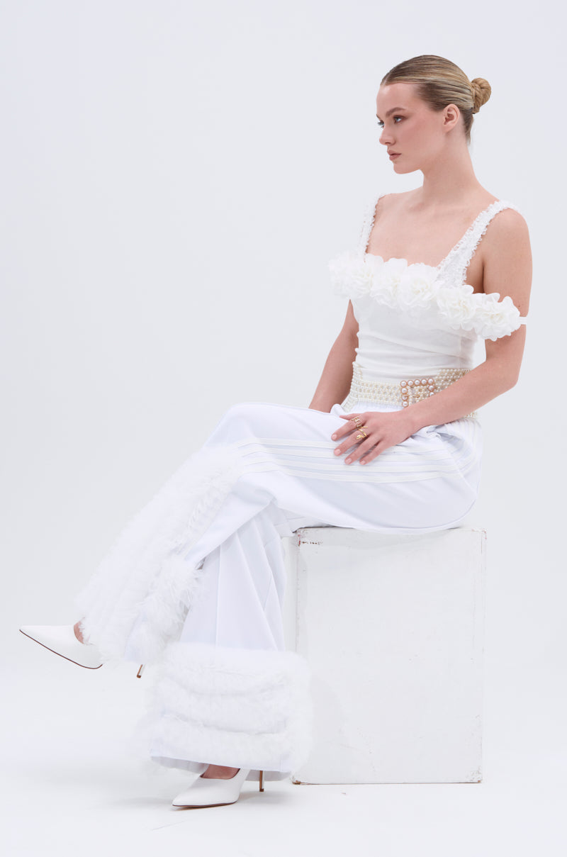 A woman with blonde hair in a sleek bun models the MODERN JAM TRACK PANT IN WHITE, featuring wide legs and furry cuffs. She sits sideways on a white block, styled with a floral white top and heels, gazing to her left.