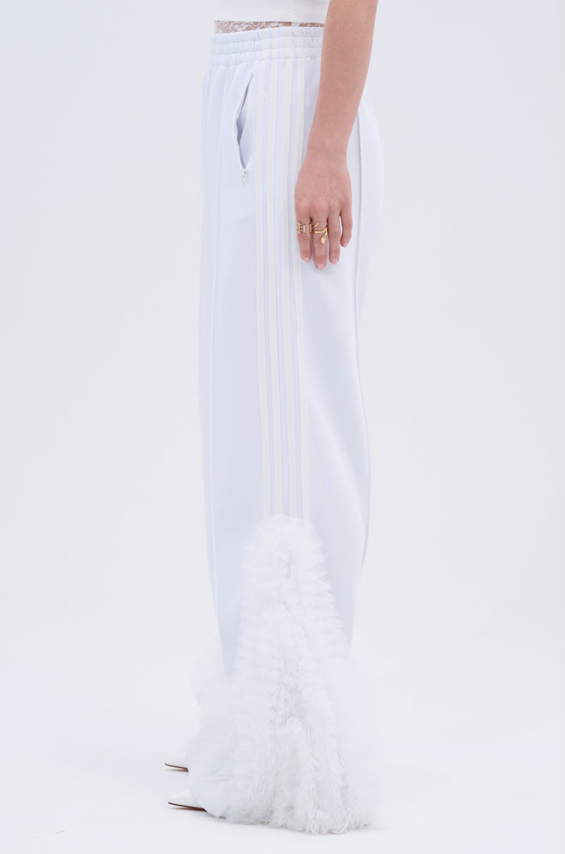 A person stands in profile wearing the MODERN JAM TRACK PANT IN WHITE, featuring high-rise fit, light side stripes, and fluffy textured fabric at the lower leg. Their arm rests by their side with rings. The background is plain and light.