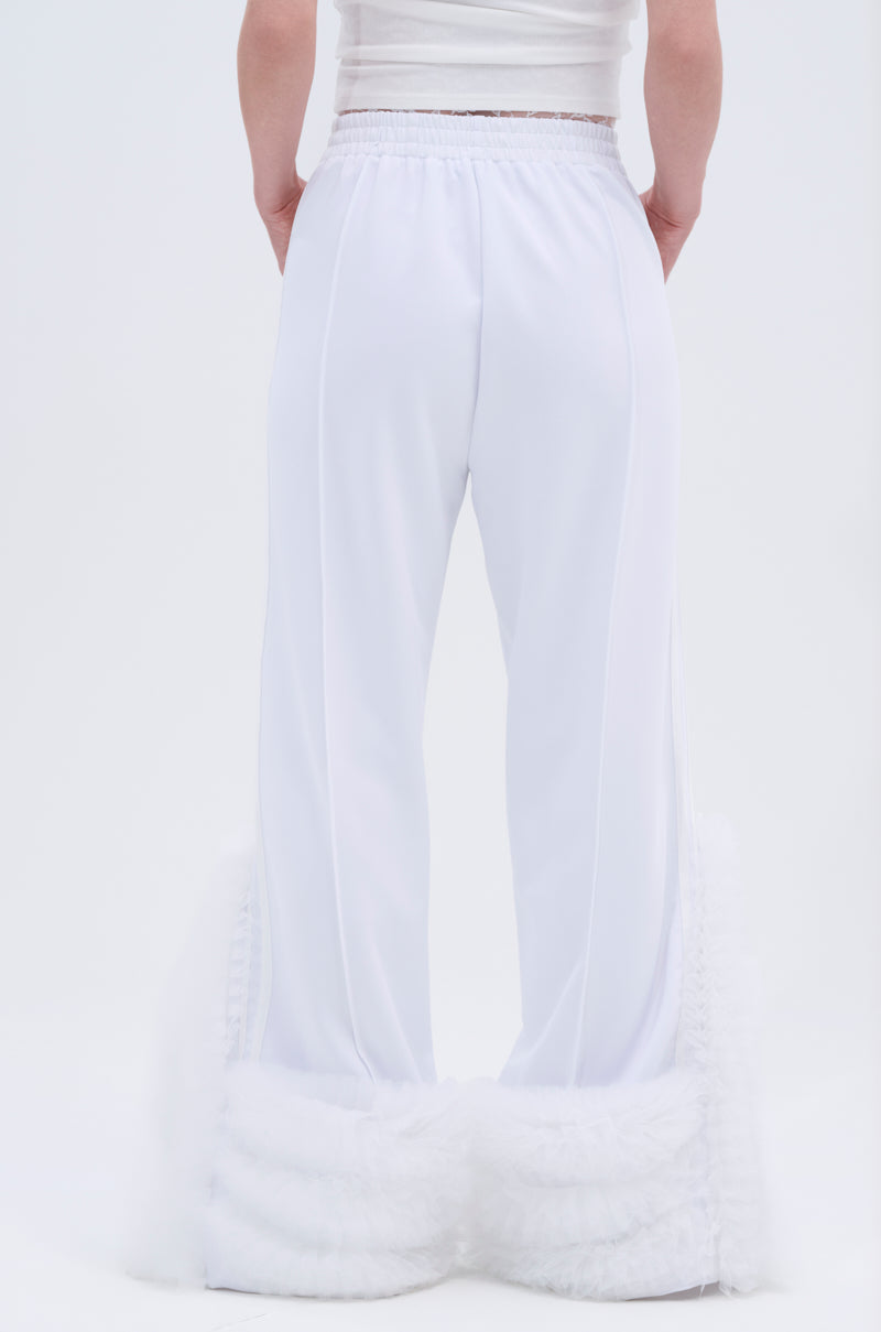 A person stands with hands behind their back, wearing the MODERN JAM TRACK PANT IN WHITE—loose-fitting, high-rise pants with ruffled sheer fabric at the bottom. The photo is taken from behind against a plain white background.