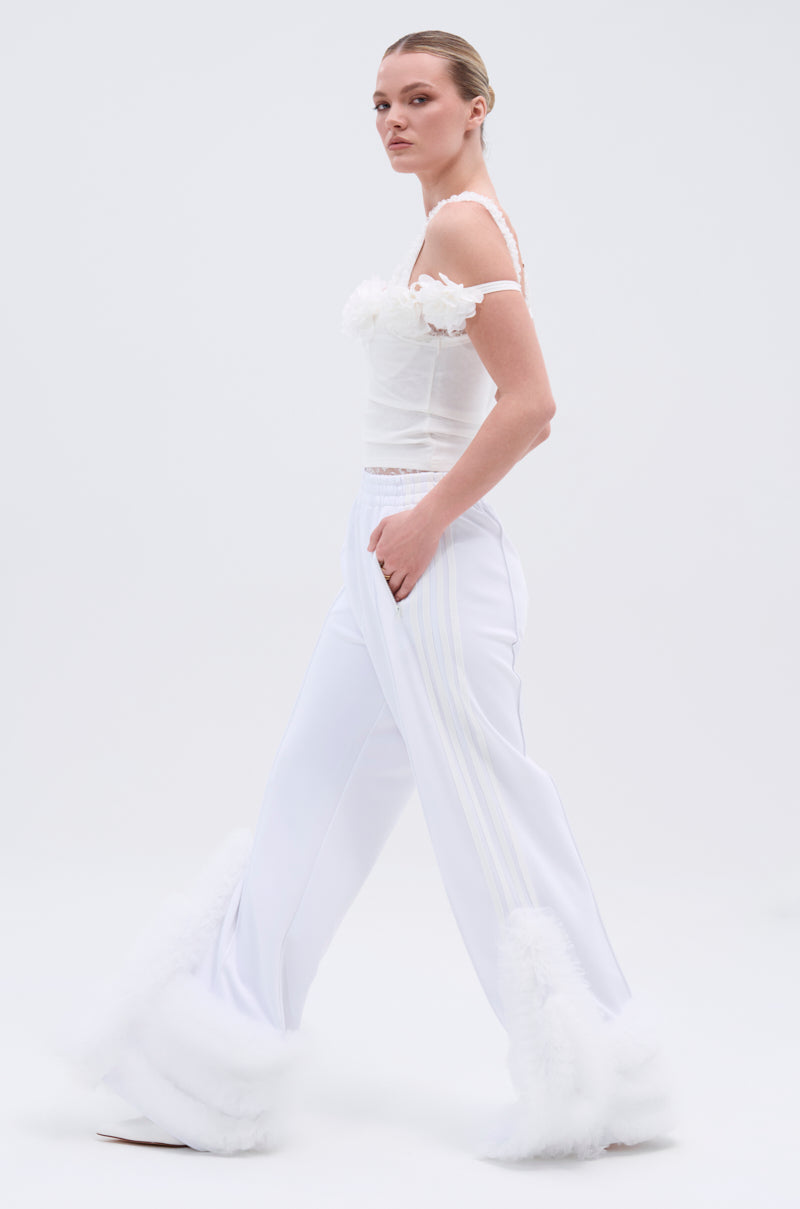 A woman walks in profile against a plain white background, wearing the MODERN JAM TRACK PANT IN WHITE—high-rise, wide-leg track pants with fluffy white hem trim—paired with a fitted white top featuring delicate straps and textured details.