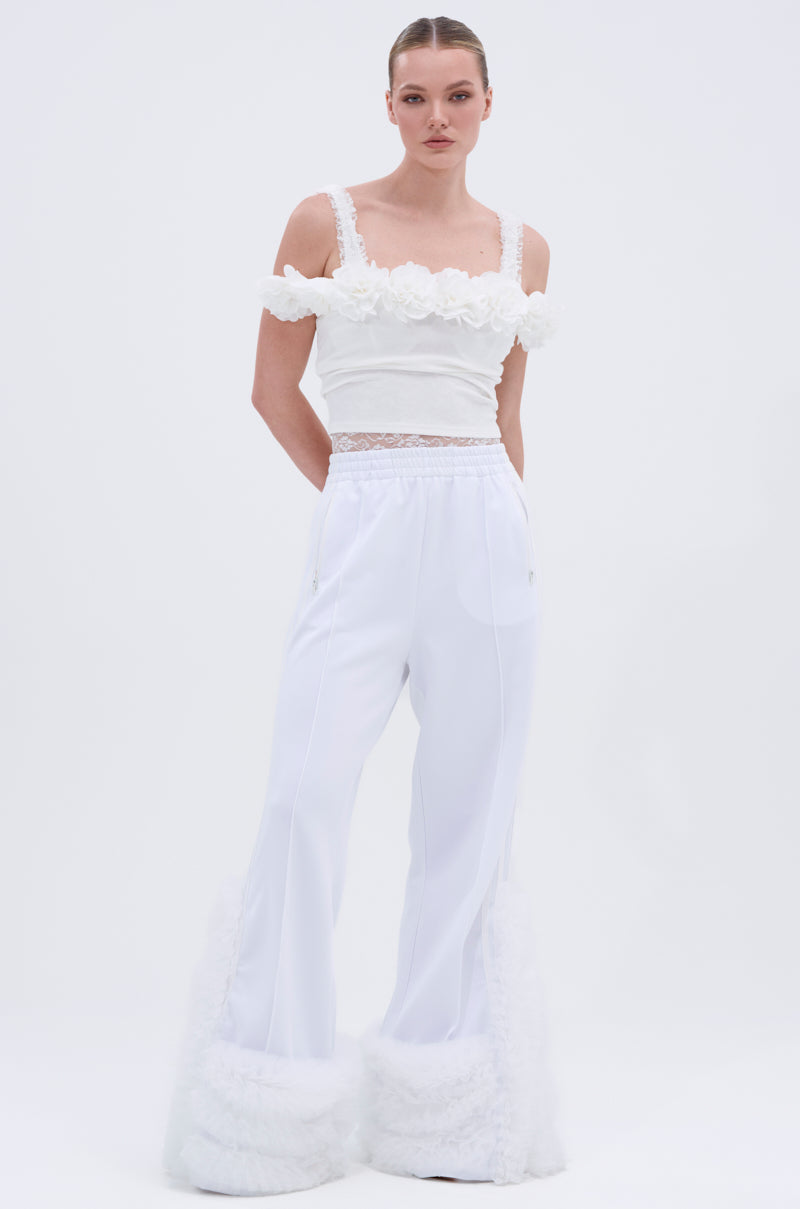 A woman stands against a plain white background, looking at the camera and wearing the MODERN JAM TRACK PANT IN WHITE paired with a sleeveless white top featuring floral details and fluffy ankle trim.