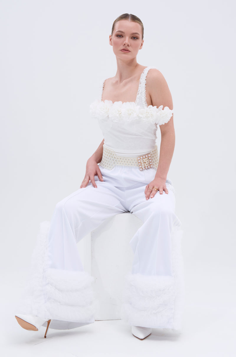 A woman wears the MODERN JAM TRACK PANT IN WHITE—high-rise, wide-leg with textured details and zippered pockets—styled with a floral-detailed top, pearl-studded belt, and white heels as she sits on a white cube against a plain background.