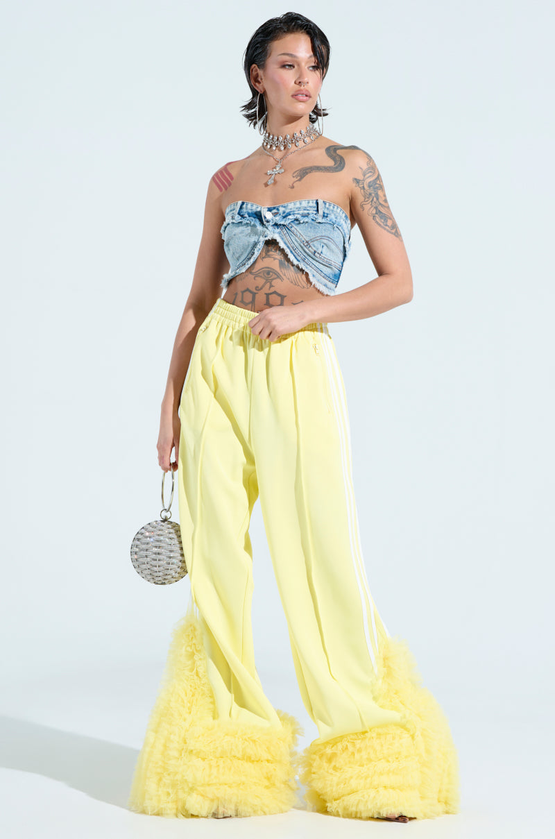 A woman poses against a light background, wearing a denim tube top and MODERN JAM TRACK PANT WITH TULLE IN YELLOW with ruffled hems. She holds a round metallic purse and is adorned with tattoos and jewelry, including a statement necklace.