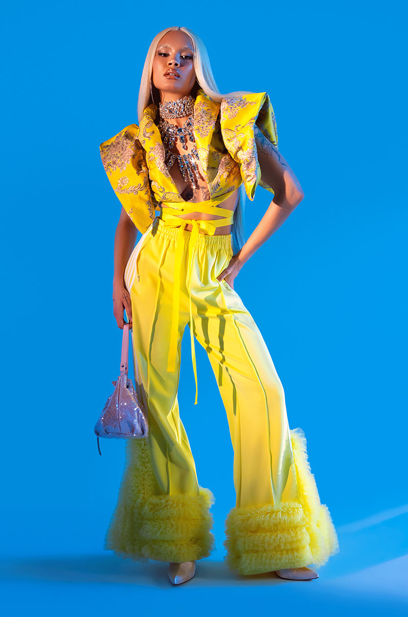 A model poses against a blue background wearing the MODERN JAM TRACK PANT WITH TULLE IN YELLOW, paired with a crystal necklace, long straight blonde hair, and a small sparkling handbag.
