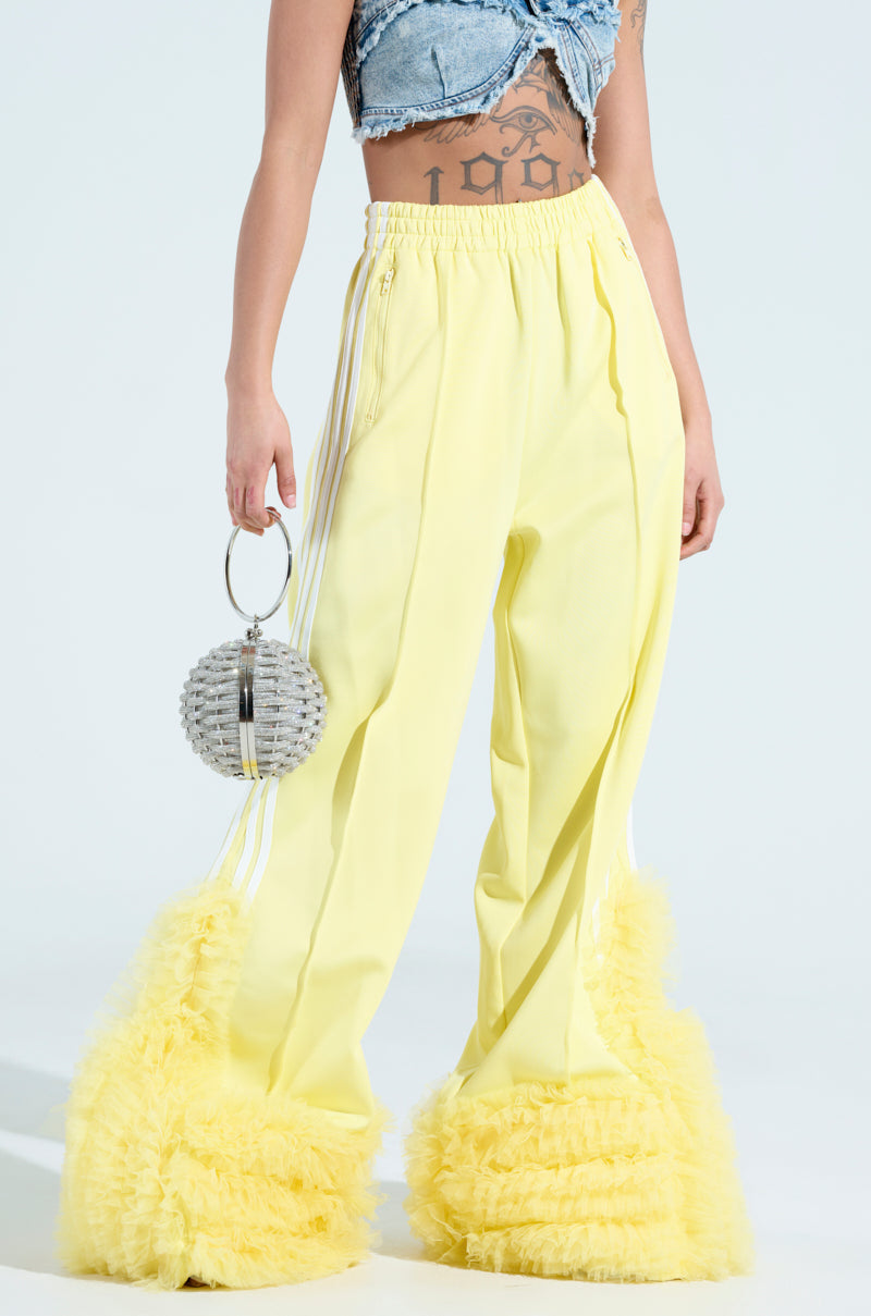 A person in MODERN JAM TRACK PANT WITH TULLE IN YELLOW, featuring high-waisted design and ruffled hems, stands holding a round purse. They're wearing a cropped, frayed denim top that reveals a geometric tattoo on their midriff against a light background.