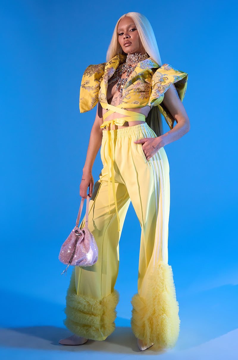 A person with long blonde hair poses confidently in the MODERN JAM TRACK PANT WITH TULLE IN YELLOW, pairing the wide-leg pants with a cropped jacket, sparkling necklace, and glittery pink handbag against a blue background.