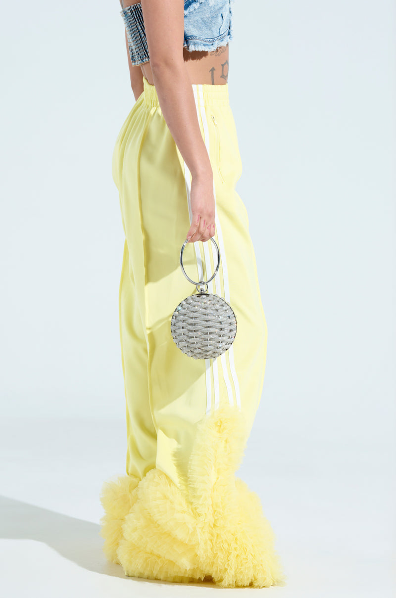 A person stands in MODERN JAM TRACK PANT WITH TULLE IN YELLOW and a blue cropped top, holding a round silver handbag with a hoop handle against a soft, light background.