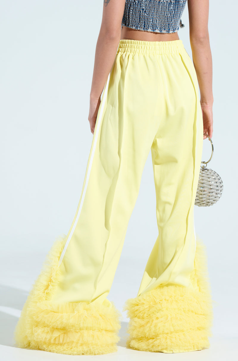 A person models the Modern Jam Track Pant with Tulle in Yellow, featuring fluffy hem ruffles and a wide-leg silhouette. They hold a small round beaded handbag and wear a sleeveless, textured blue crop top against a plain light blue background.