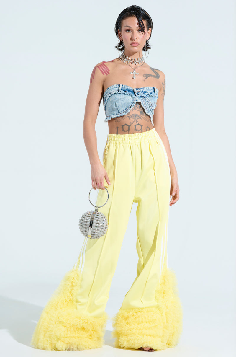 A person poses against a plain background, wearing a denim bandeau top and MODERN JAM TRACK PANT WITH TULLE IN YELLOW. They hold a spherical handbag, sport short wet-look hair, and display tattoos on their midriff and arms, complementing the wide-leg silhouette of their ensemble.