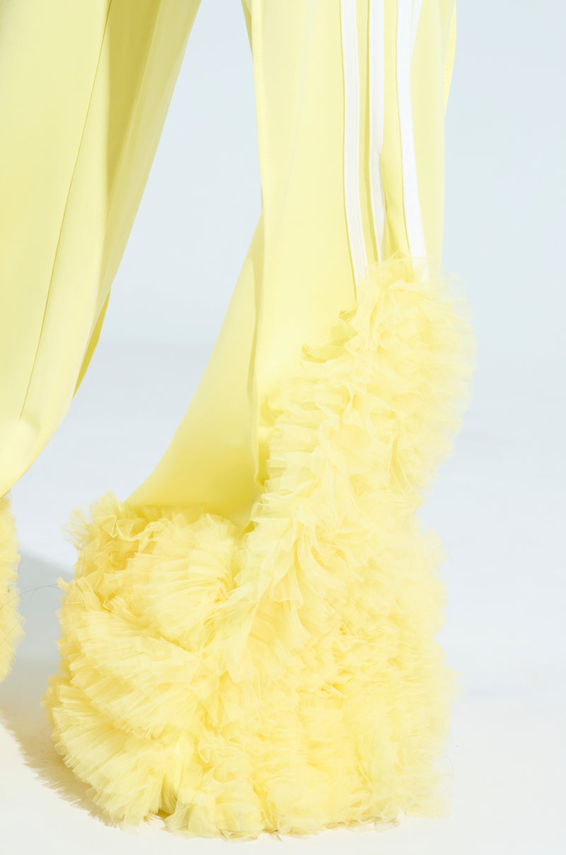 The MODERN JAM TRACK PANT WITH TULLE IN YELLOW features cascading ruffles at the hem, offering a fluffy, textured look. Its wide-leg silhouette and lightweight fabric contribute to a soft, airy feel in the design.