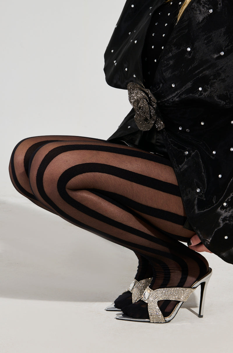 A person dressed in a black velvet outfit with pearl embellishments kneels, highlighting the OH YES STRIPE TIGHTS. They complement their look with heeled sandals decorated with sparkling bows.