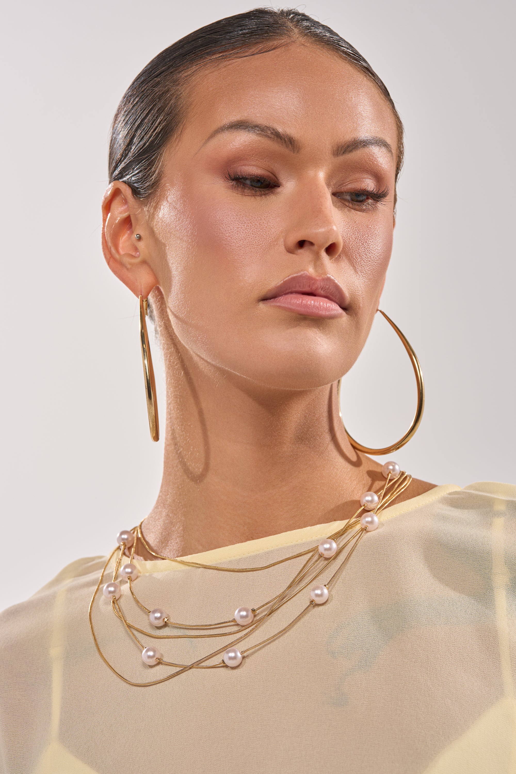 A woman with slicked-back hair wears large gold hoops, a sheer cream top, and the SHES EXPENSIVE NECKLACE—a bold gold-tone chain with pearls—posing against a plain light background. 1