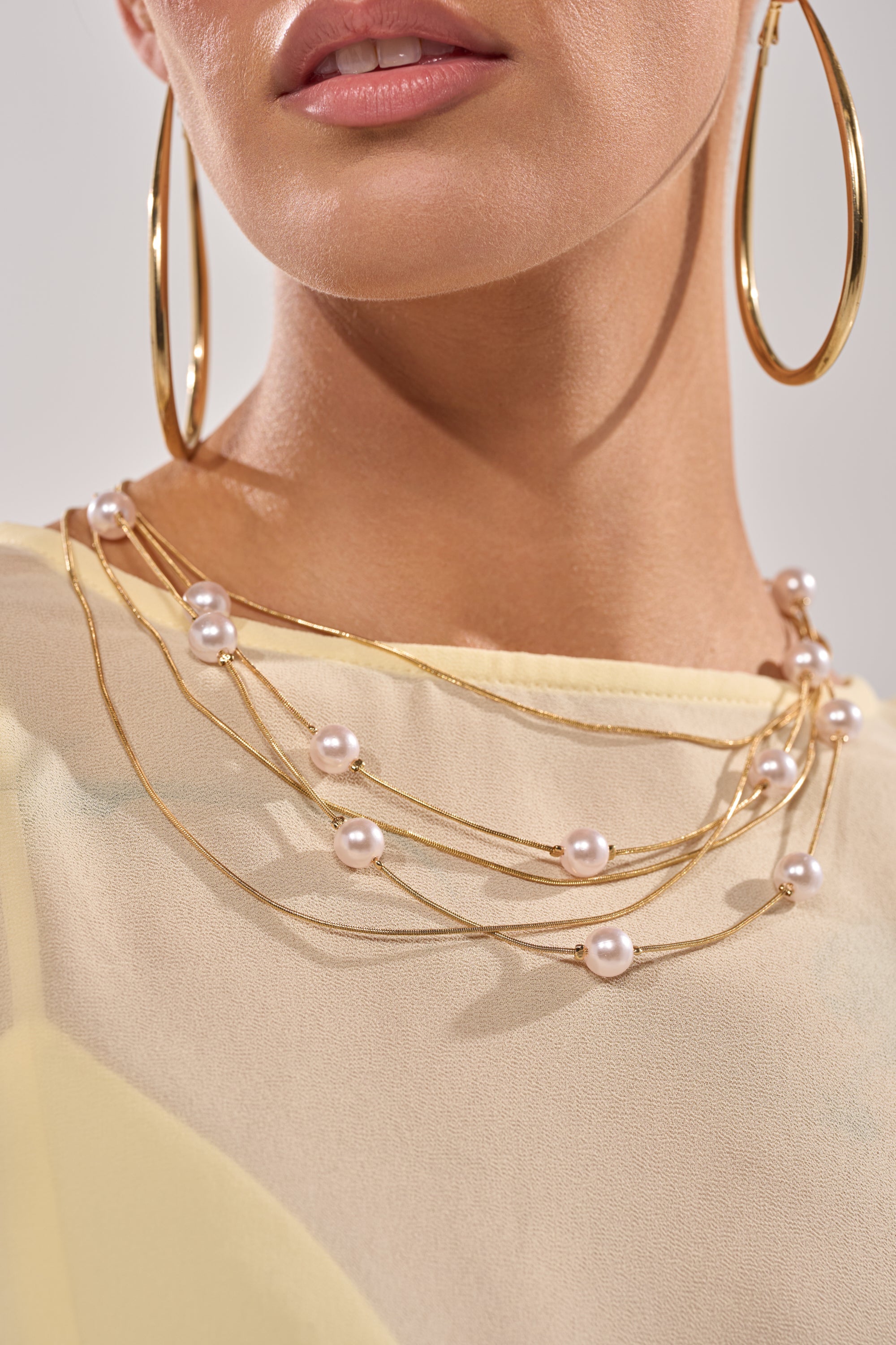 A woman in a light yellow top wears large gold hoops and the SHES EXPENSIVE NECKLACE—a layered pearl necklace with gold accents. 2