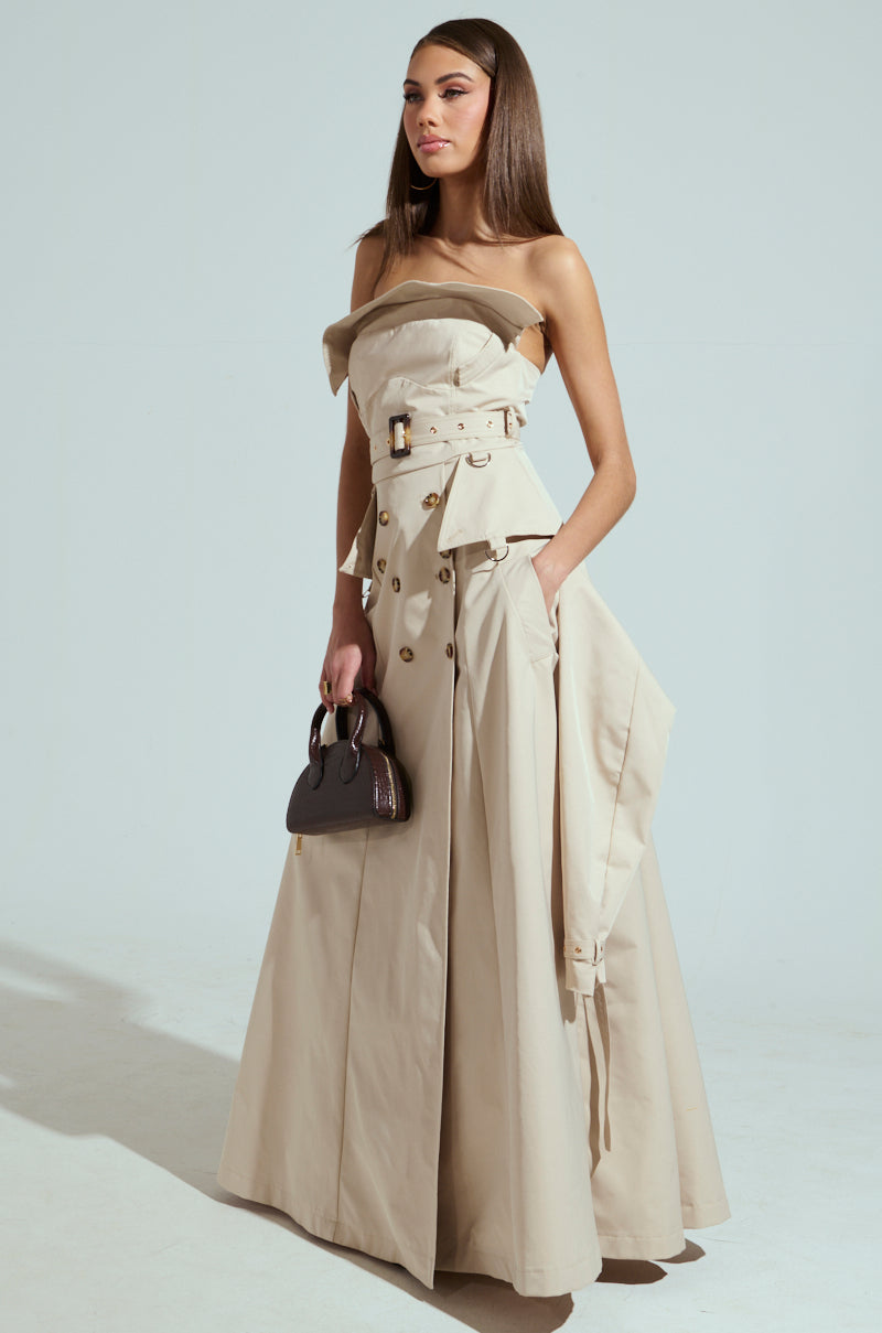 A woman stands confidently in a floor-length, strapless beige trench maxi dress with a belted waist and buttons. She's holding a small black handbag, and her long straight hair cascades down. The scene is set against a simple light gray background.