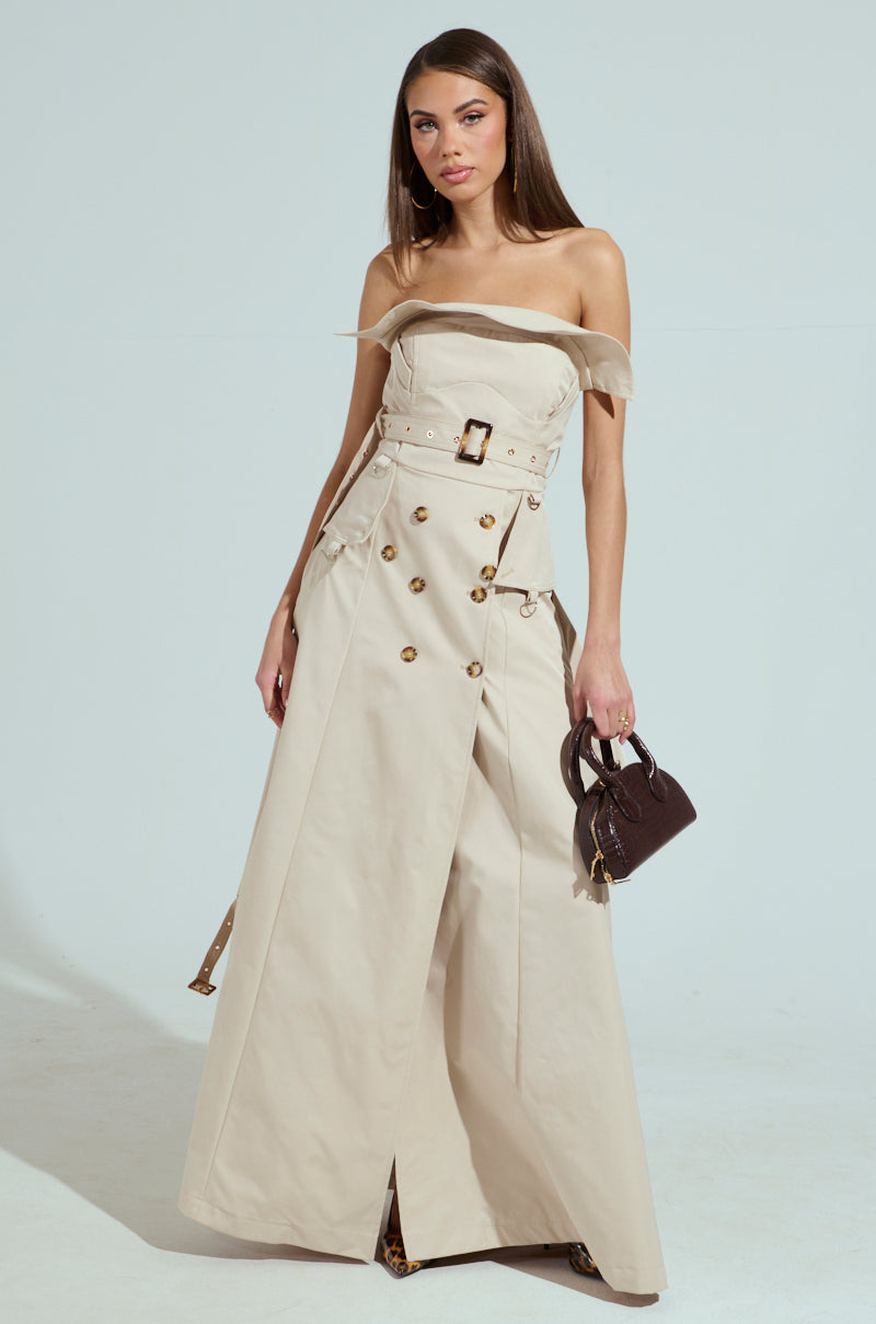 A woman poses in a STRAPLESS TRENCH MAXI DRESS, styled with buttons and a belt. She accessorizes with a small brown handbag and hoop earrings. The dress features a flared, floor-length skirt, creating an elegant look against the plain light gray background.