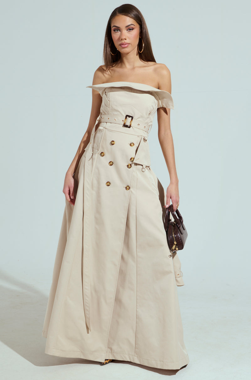A woman elegantly wears a strapless trench maxi dress, clutching a small dark handbag. She stands poised against a plain background, exuding sophistication.