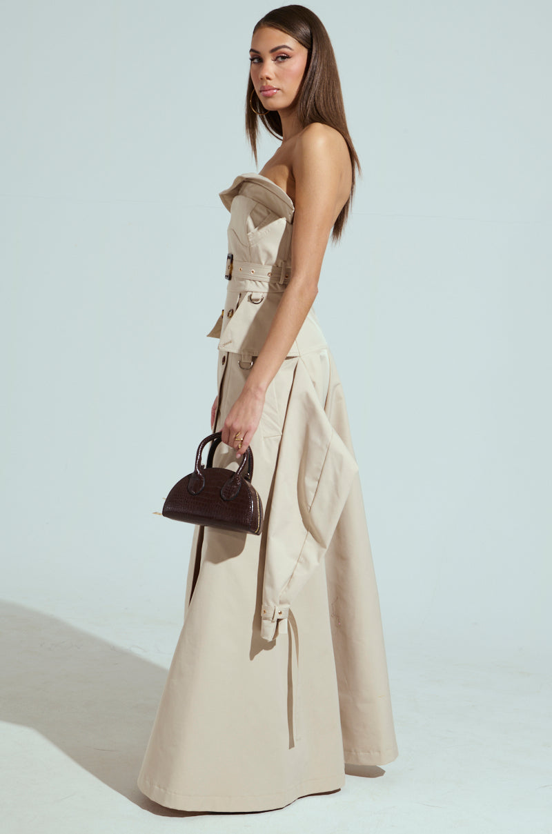 A woman stands confidently in a beige STRAPLESS TRENCH MAXI DRESS, with a matching coat draped over one arm. She holds a small brown handbag, and her long hair is sleek and straight against the plain light gray background.