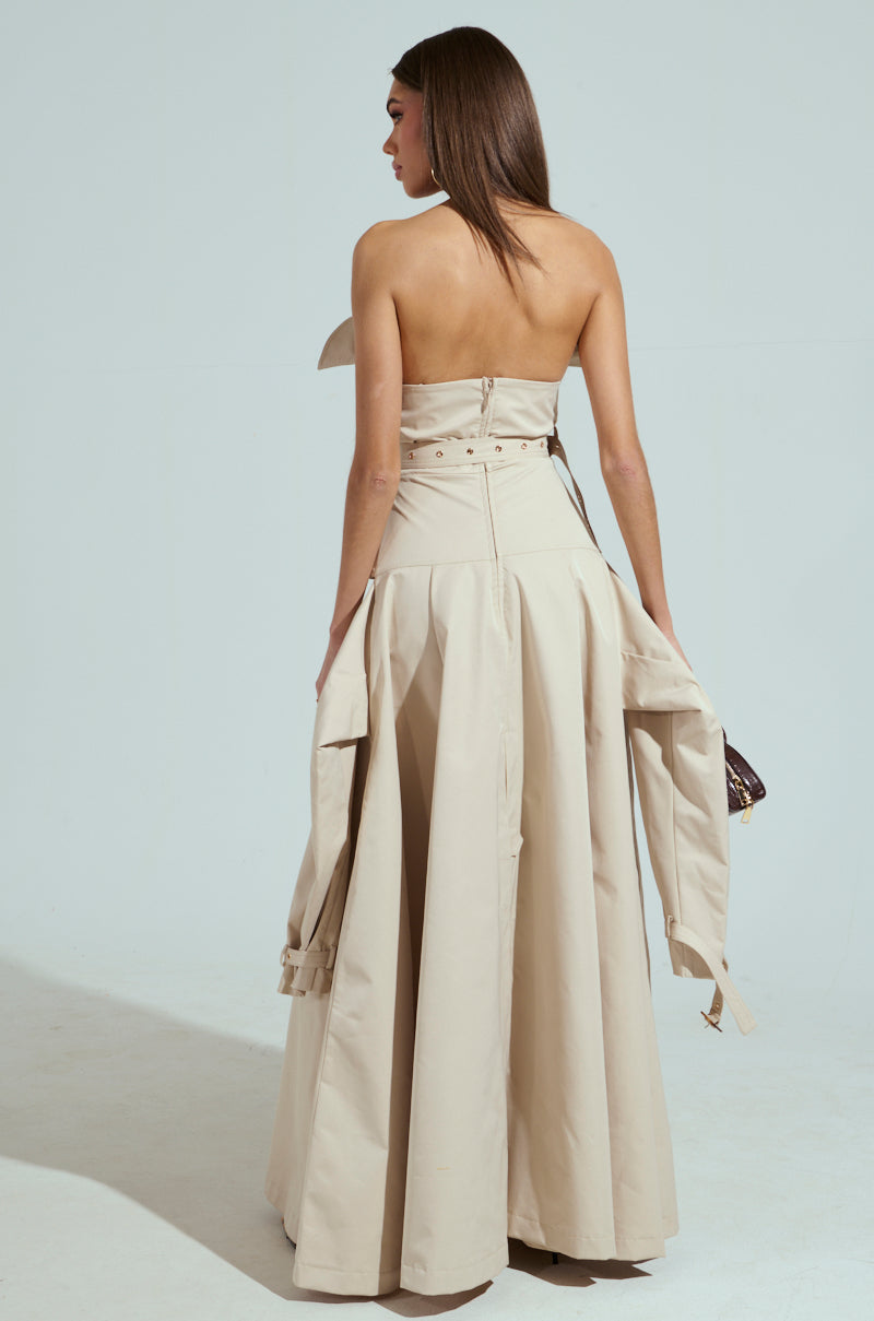 A woman, with straight brown hair draped over her shoulders, faces away from the camera wearing a STRAPLESS TRENCH MAXI DRESS. The beige gown has a long, flowing skirt and a jacket hanging loosely from its belted waist against a plain light gray background.