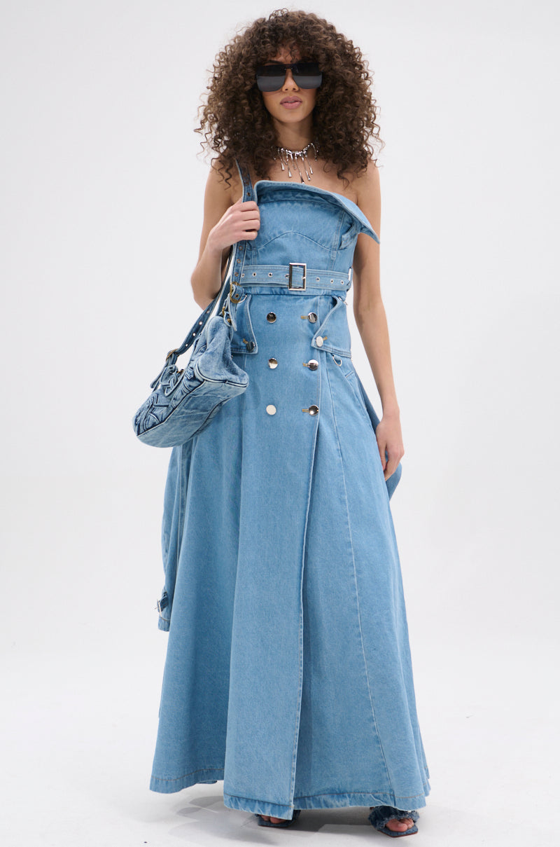 A woman with curly hair wears the STRAPLESS TRENCH MAXI DRESS IN DENIM, oversized sunglasses, a chunky necklace, and carries a matching denim handbag while standing against a plain white background.