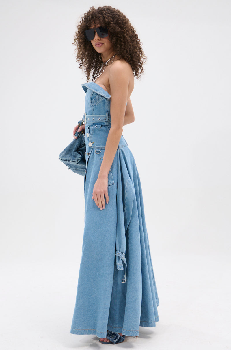 A woman with curly hair wears the STRAPLESS TRENCH MAXI DRESS IN DENIM, paired with dark sunglasses and a matching denim bag, posing confidently against a plain white background.