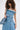 A woman with curly hair wears oversized sunglasses, a silver necklace, and the STRAPLESS TRENCH MAXI DRESS IN DENIM with double-breasted buttons, completing her look with a matching denim bag over her shoulder.