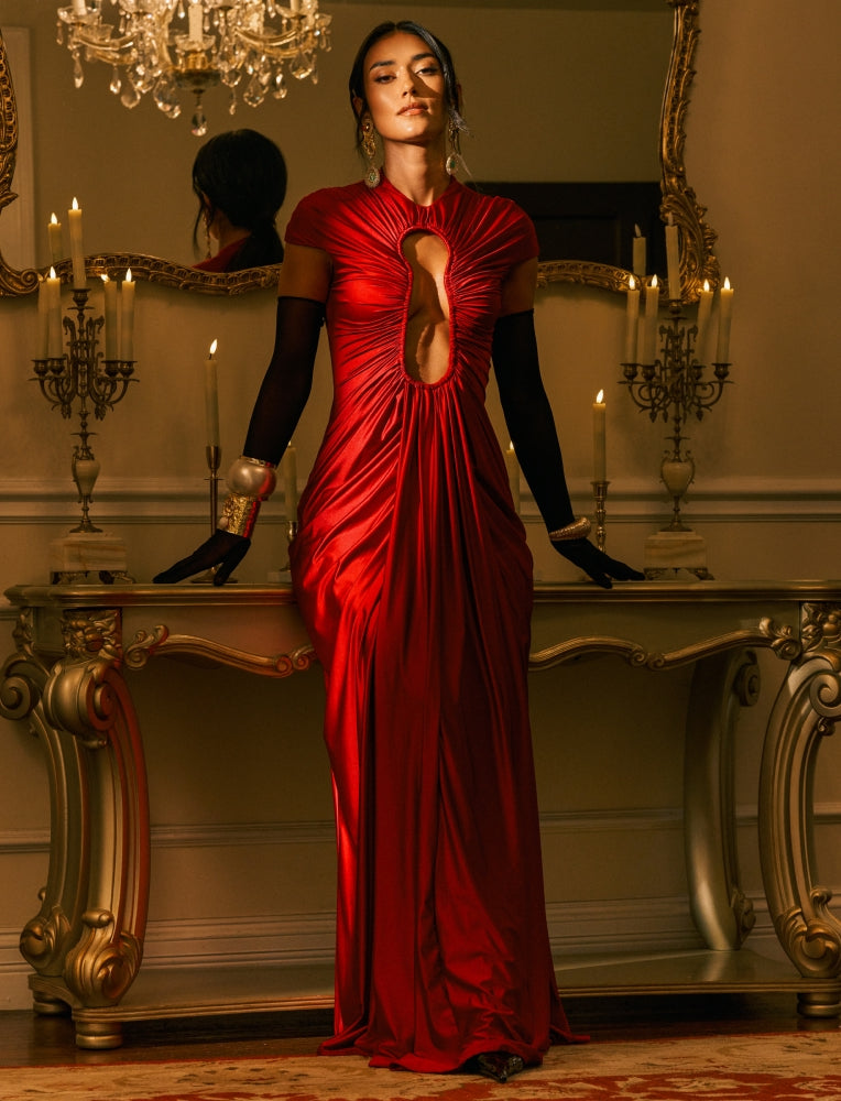 A woman stands in a luxurious room wearing a floor-length red gown with dramatic draped folds, a central cutout, and black gloves. She is posed behind an ornate gold console table with lit candles and a mirror reflecting her profile.
