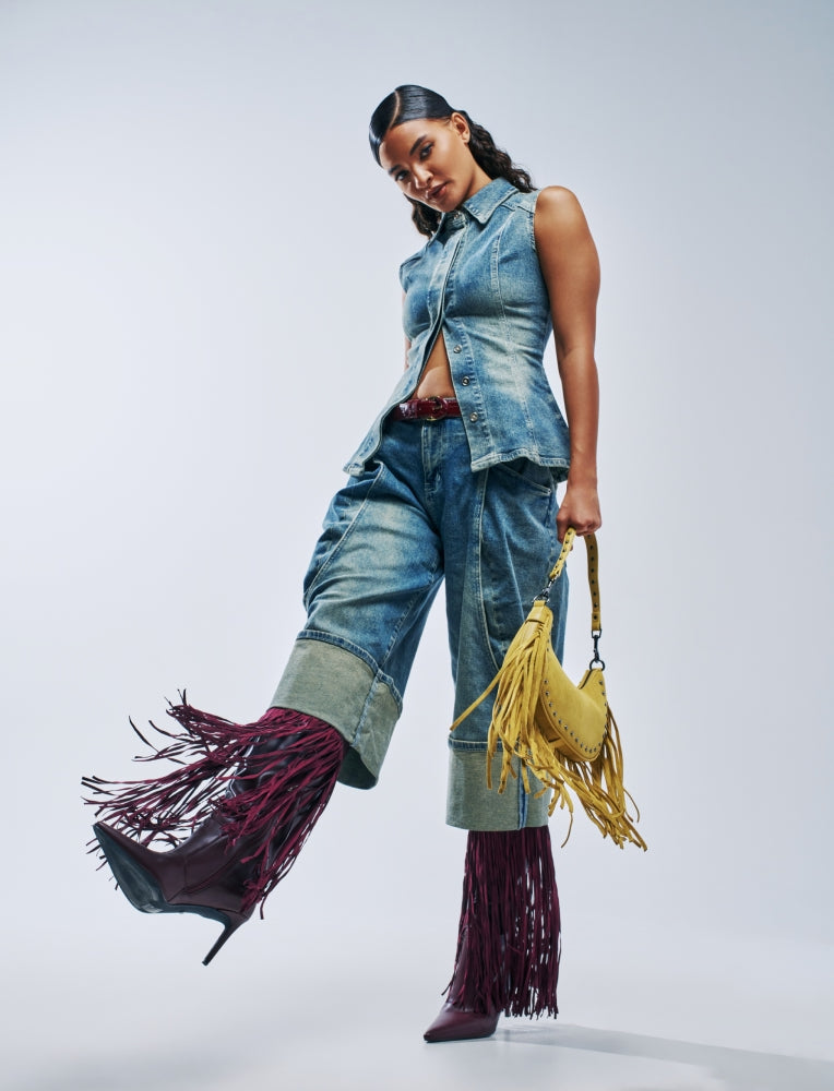 A model poses in a denim sleeveless vest and matching denim cropped pants, styled with dark burgundy fringe boots and a yellow fringe handbag against a light gray studio background.