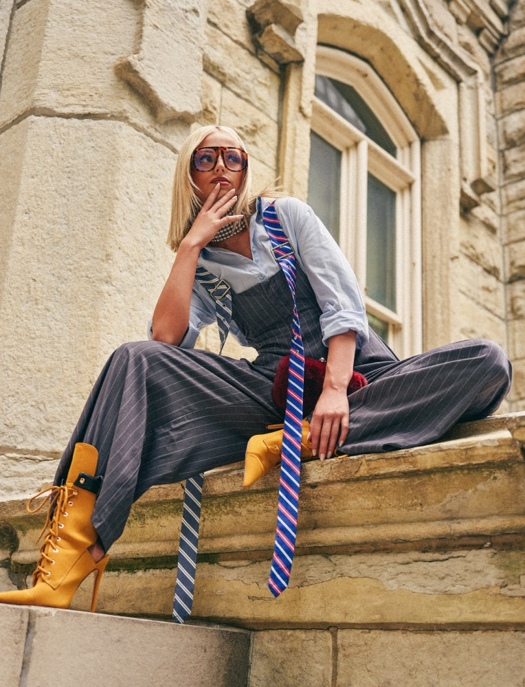 A woman sits on stone steps in front of a stone building, wearing a light blue shirt, dark pinstripe overalls, a blue and red striped tie, yellow lace-up ankle boots, and sunglasses.