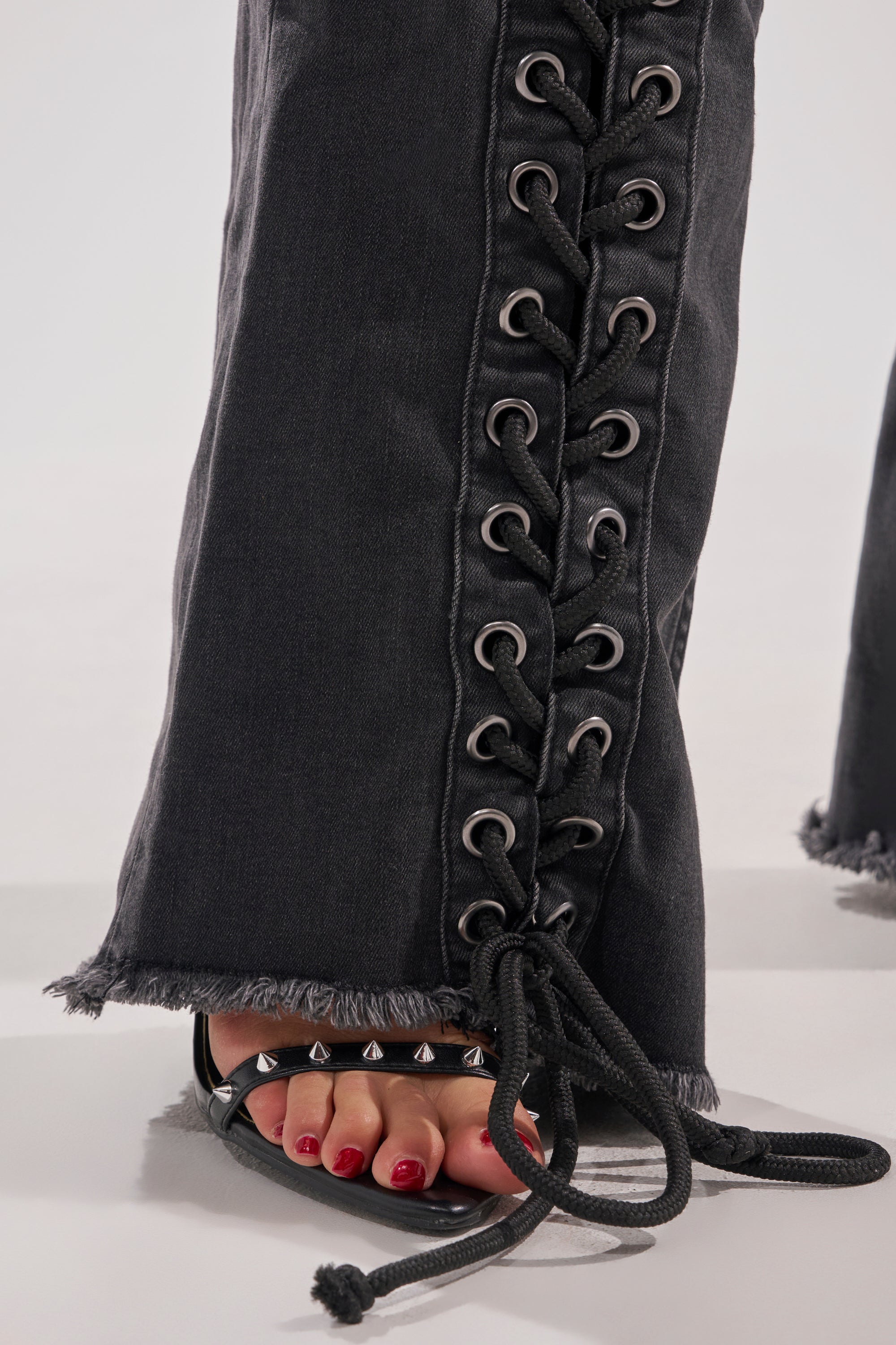 A close-up of a person's lower legs shows them wearing HOTTY LACE UP FLARE DENIM with eyelet details and frayed hems, styled with black spiked sandals and red painted toenails. 9