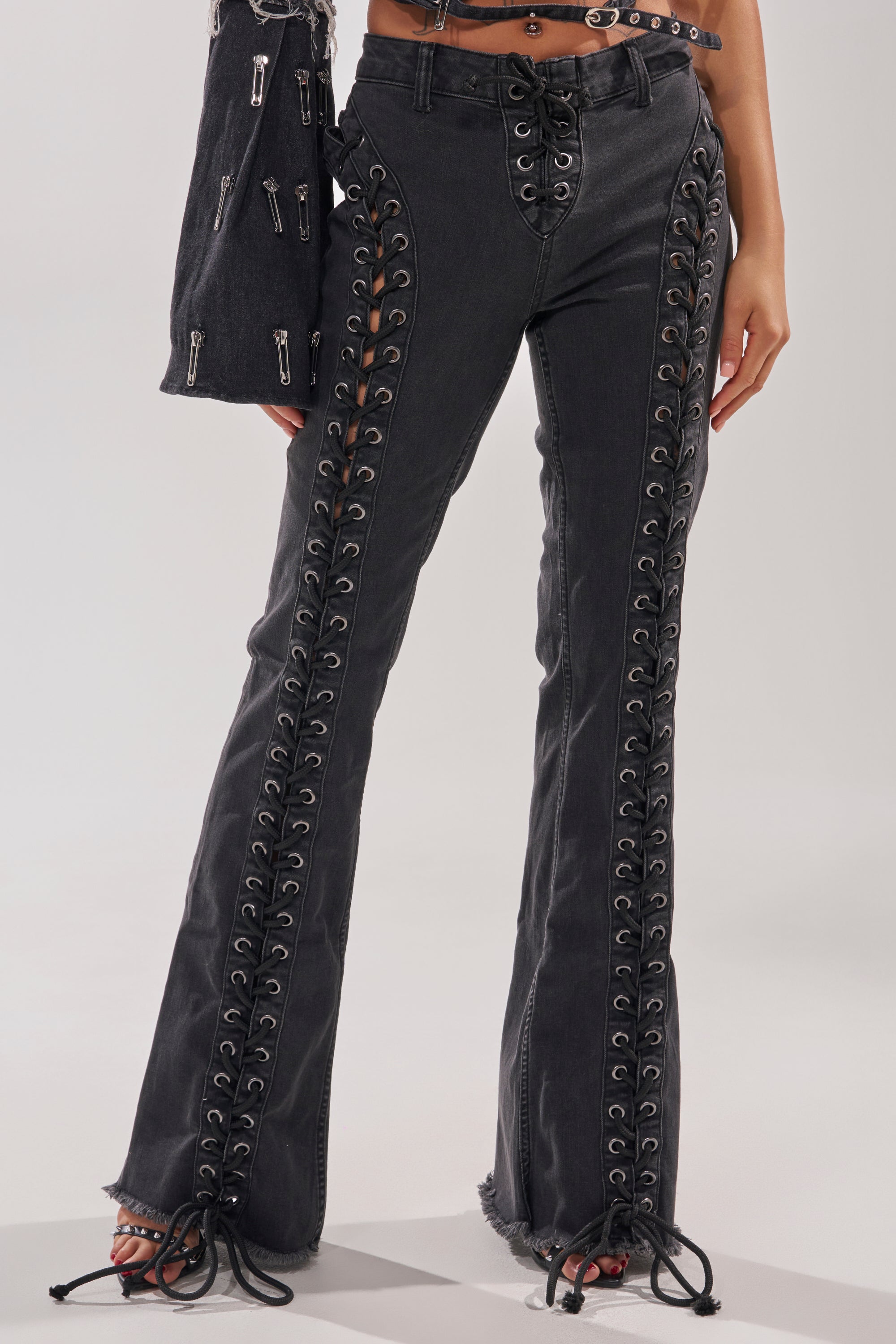 Wearing the HOTTY LACE UP FLARE DENIM—curve-hugging black flare jeans with bold grommets and side lace-ups revealing skin—a person holds a matching black garment. 1