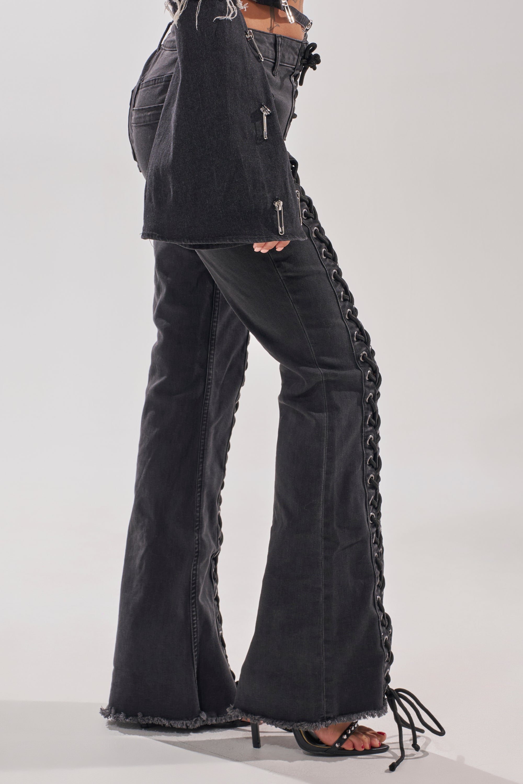 Model wears the HOTTY LACE UP FLARE DENIM with bold side lace-up details, styled with a matching black jacket and heeled sandals, standing against a light-colored background. 2