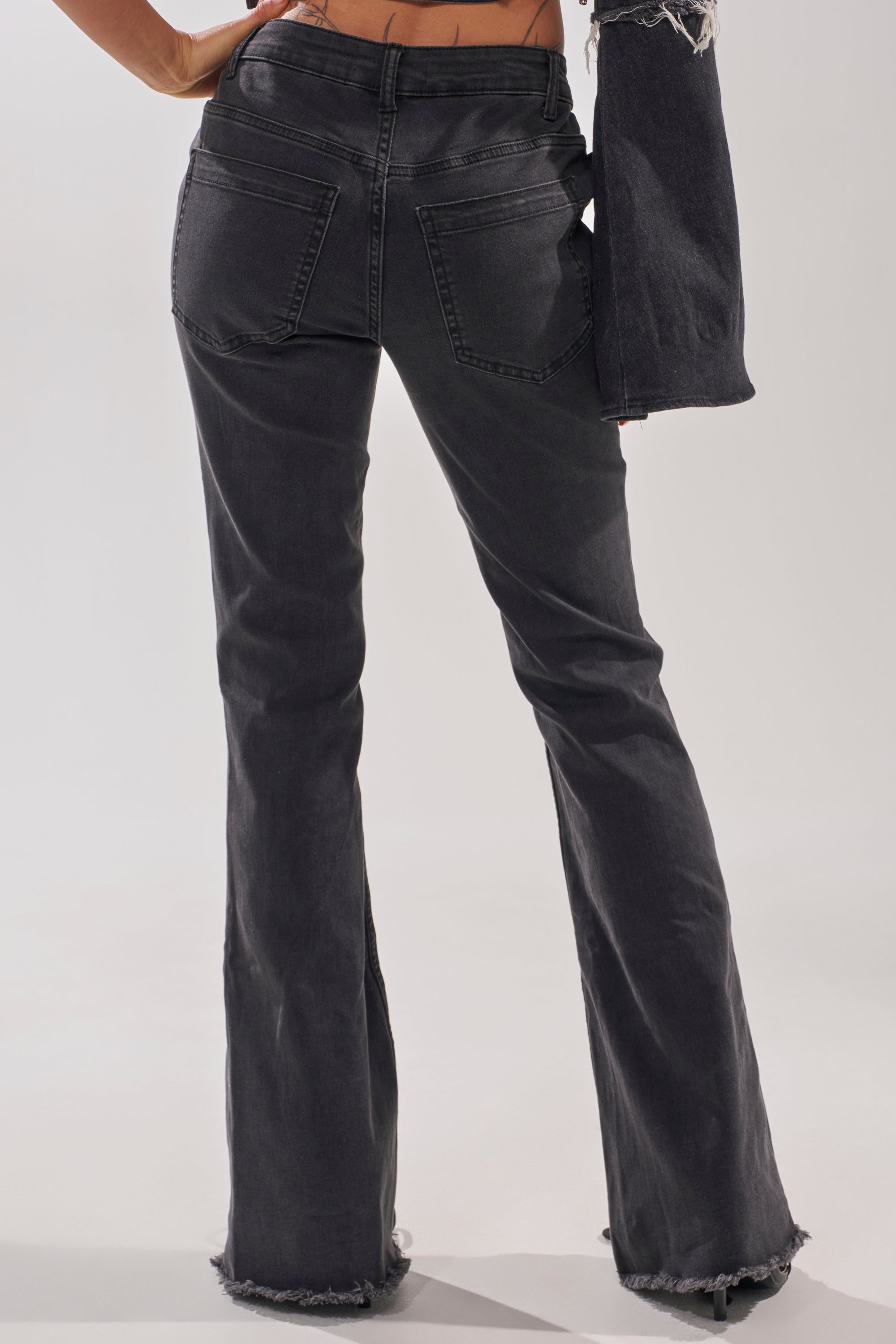 A person wears HOTTY LACE UP FLARE DENIM jeans with frayed hems, seen from the back; their left hand is on their hip and right arm at their side. 3