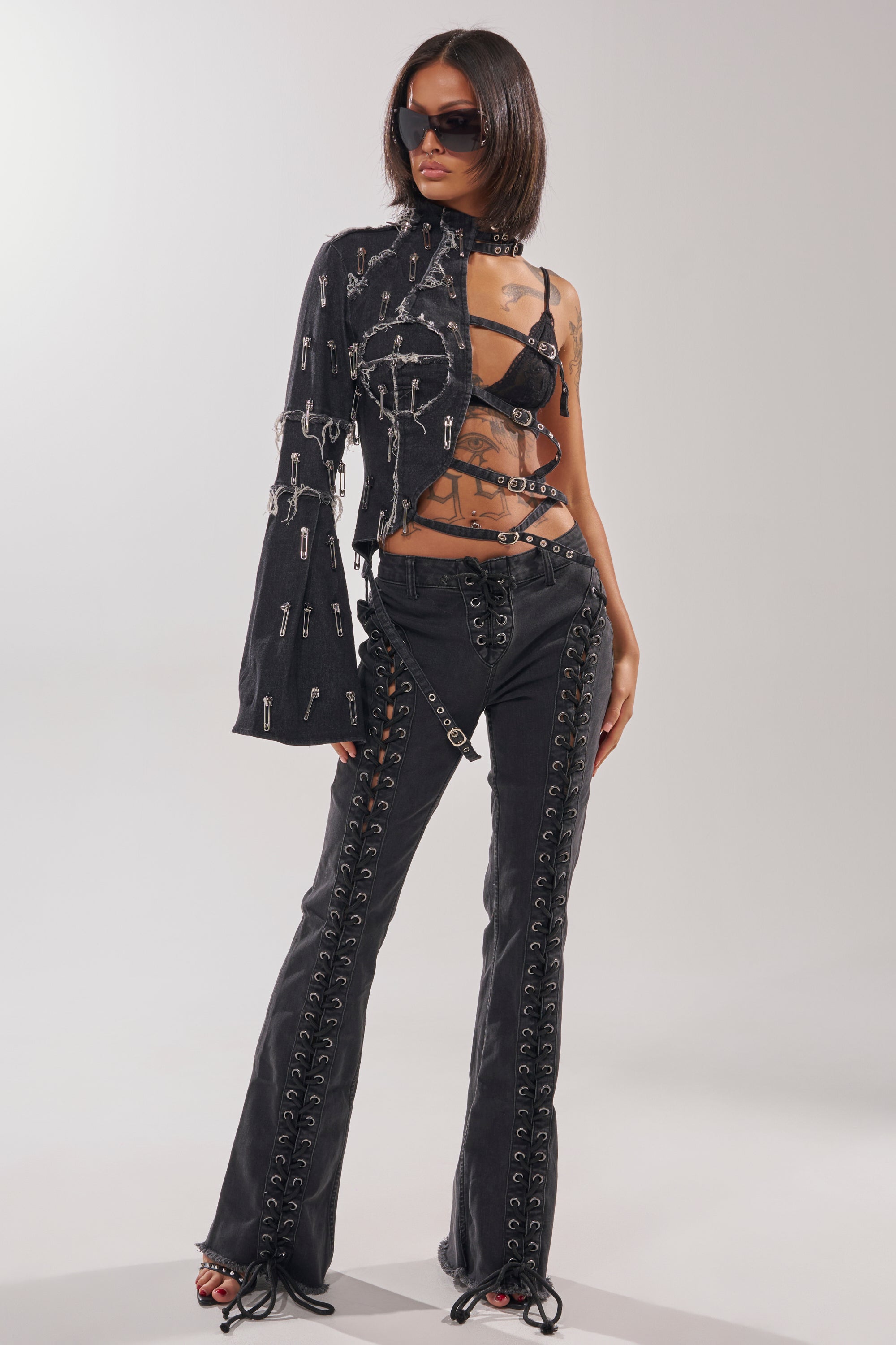 A woman confidently poses against a plain background wearing HOTTY LACE UP FLARE DENIM, a strappy black top, a safety pin-embellished one-sleeve jacket, black strappy heels, and sunglasses. 6