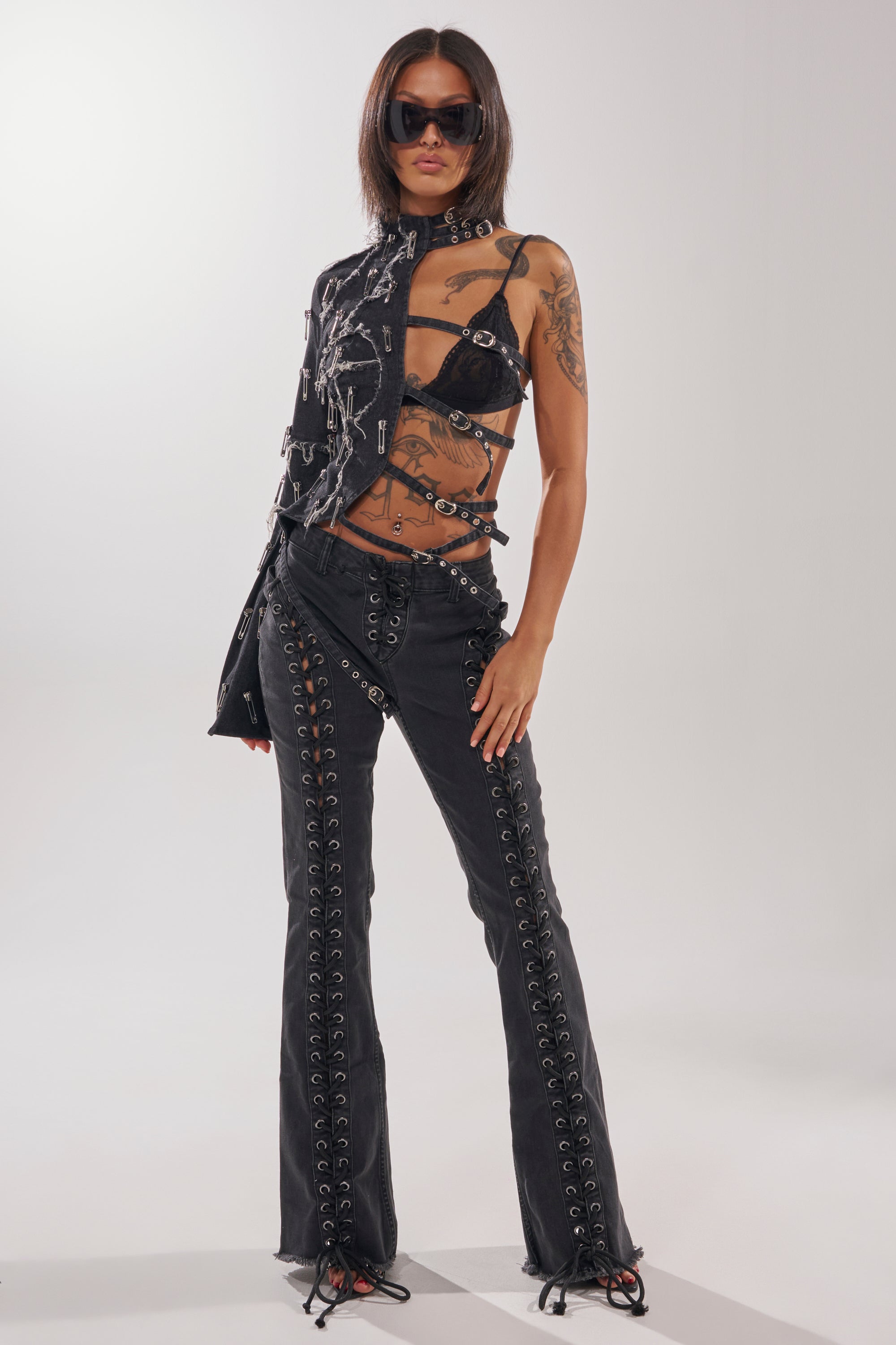 Model wears the HOTTY LACE UP FLARE DENIM, paired with an asymmetrical black mesh top featuring straps and metal rings, black sunglasses, and displays visible tattoos against a plain light background. 7