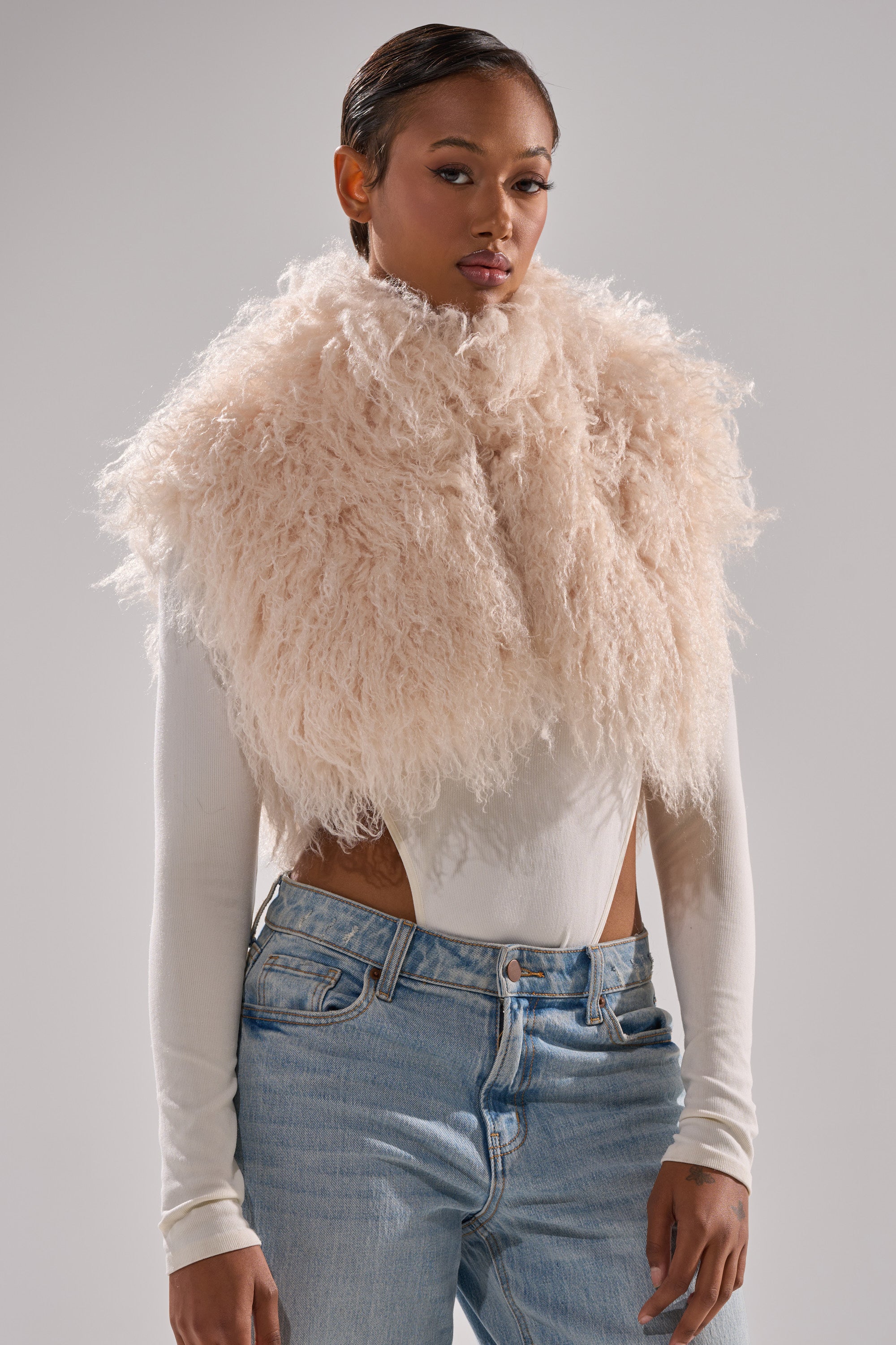 A woman models the COCO CROPPED FUR VEST IN BEIGE over a long-sleeve white top and light blue jeans, posing against a plain gray background.