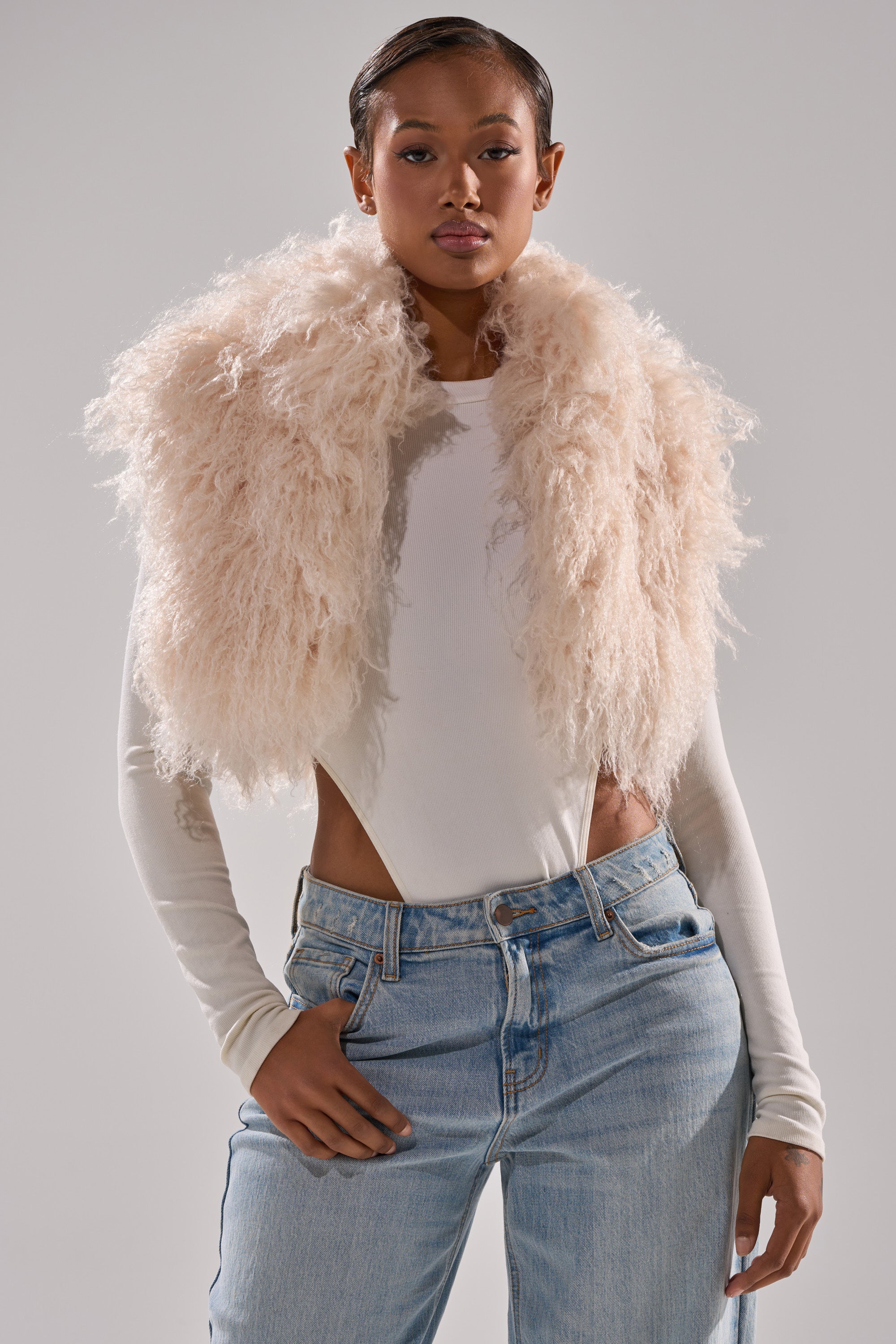 A woman poses confidently in a long-sleeve white bodysuit, light blue jeans, and the COCO CROPPED FUR VEST IN BEIGE draped over her shoulders against a plain gray background.